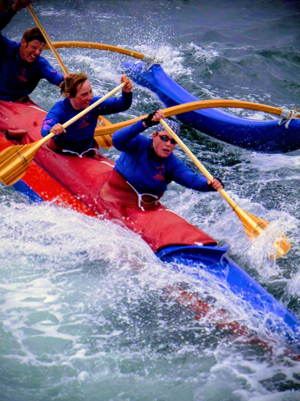 Drei Paddler in einem Outrigger-Kanu kämpfen sich durch starke Wellen und Gischt, Fokus auf die dynamische Paddelbewegung.