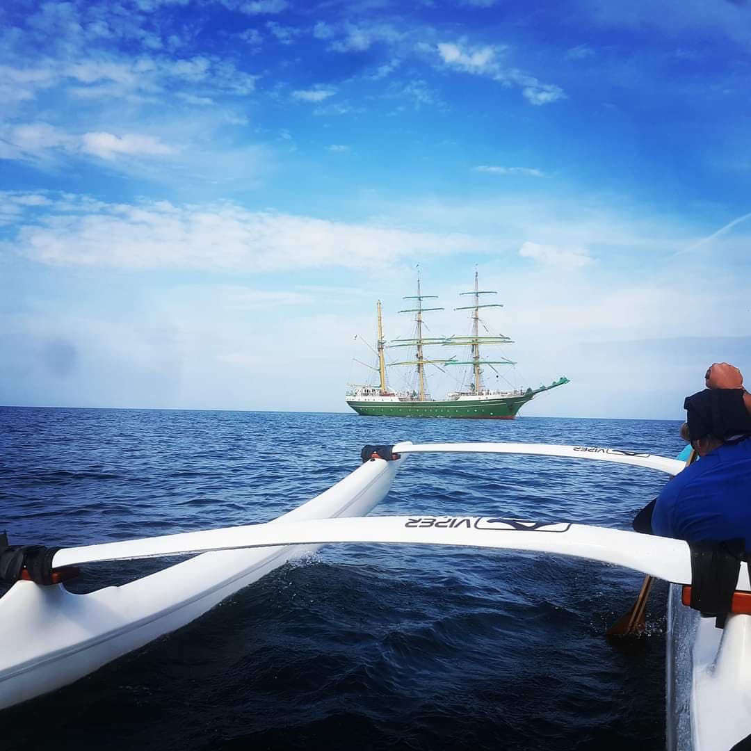 Perspektive aus einem weißen Outrigger-Kanu auf ein großes, grünes Dreimast-Segelschiff auf offener See unter blauem Himmel.